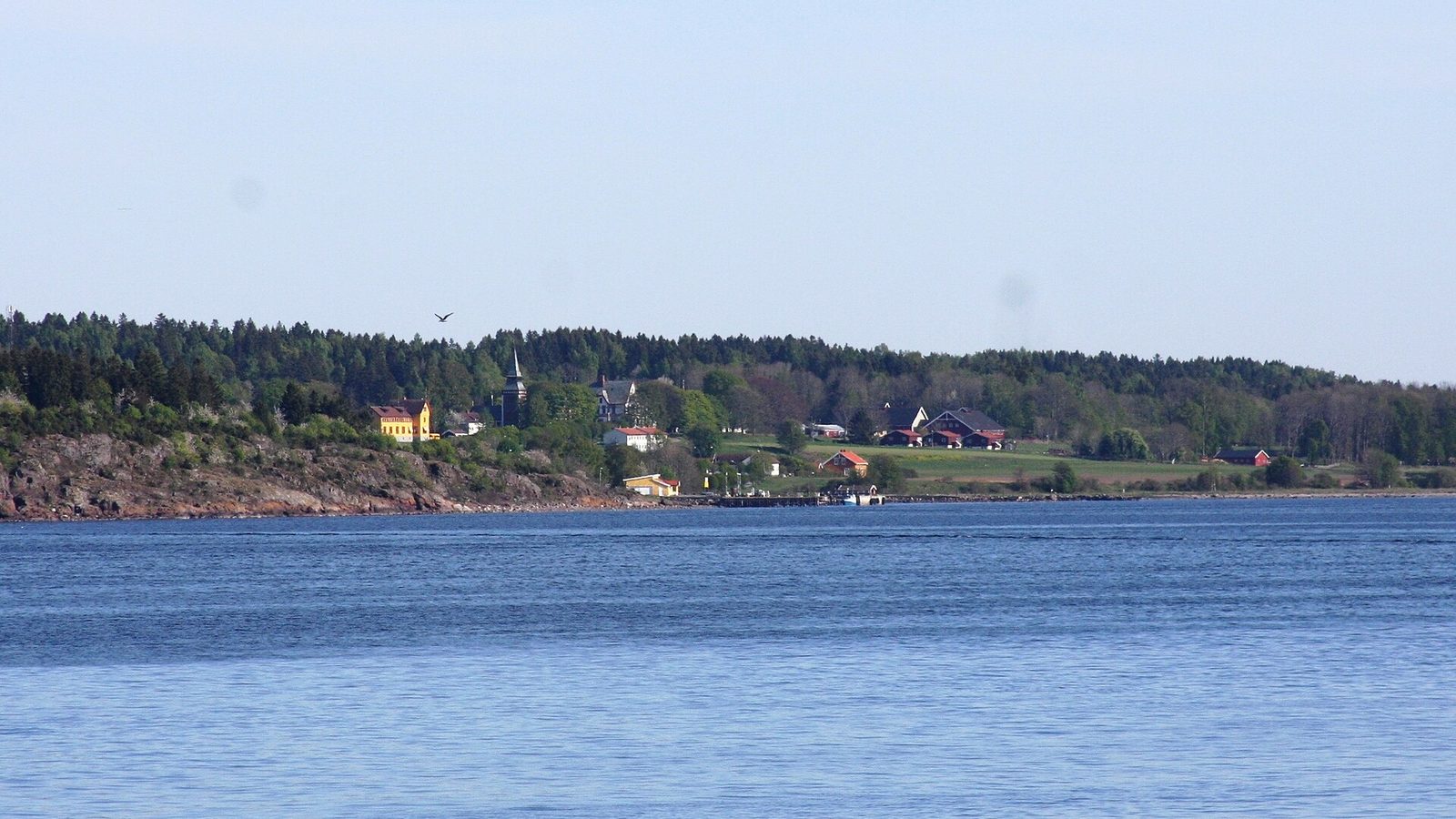 Wyspa Bastøy widziana od strony fiordu – las, żółty budynek i zabudowania gospodarstwa nad wodą